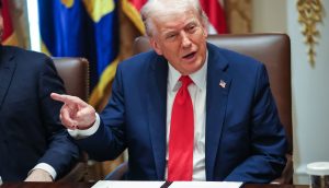 Der Mann, der mit einem Post die Kapitalmärkte zum Einsturz bringt Donald Trump in dunklem Anzug und roter Krawatte, der Trump ähnelt, sitzt an einem Tisch mit Dokumenten und gestikuliert mit seiner linken Hand, während im Hintergrund Fahnen und Papiere zu sehen sind.