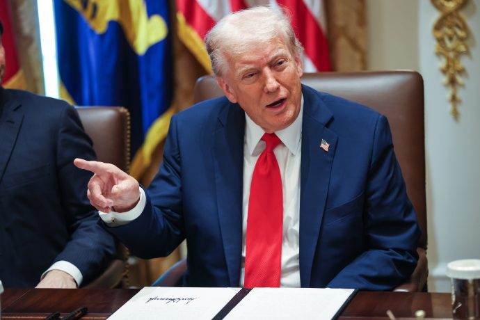 Donald Trump in dunklem Anzug und roter Krawatte, der Trump ähnelt, sitzt an einem Tisch mit Dokumenten und gestikuliert mit seiner linken Hand, während im Hintergrund Fahnen und Papiere zu sehen sind.