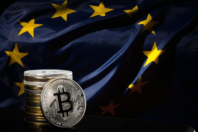 Gestapelte silberne Bitcoin-Münzen befinden sich vor einer Flagge der Europäischen Union mit sichtbaren gelben Sternen auf dunkelblauem Hintergrund.