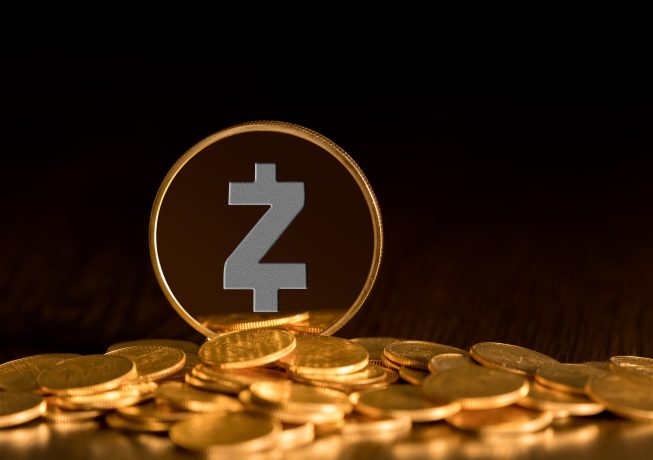 Eine physische Darstellung einer Zcash-Münze, der führenden privaten Münze, steht aufrecht zwischen verstreuten Goldmünzen auf einer dunklen Fläche.