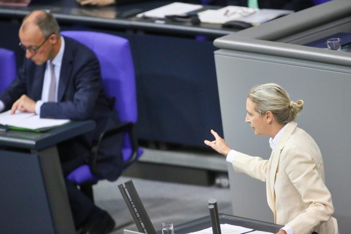 AfD-Chefin Alice Weidel in einem hellen Anzug spricht und gestikuliert an einem Podium, während Bundeskanzler Merz in einem dunklen Anzug an einem nahe gelegenen Schreibtisch sitzt und in einem formellen Rahmen liest.