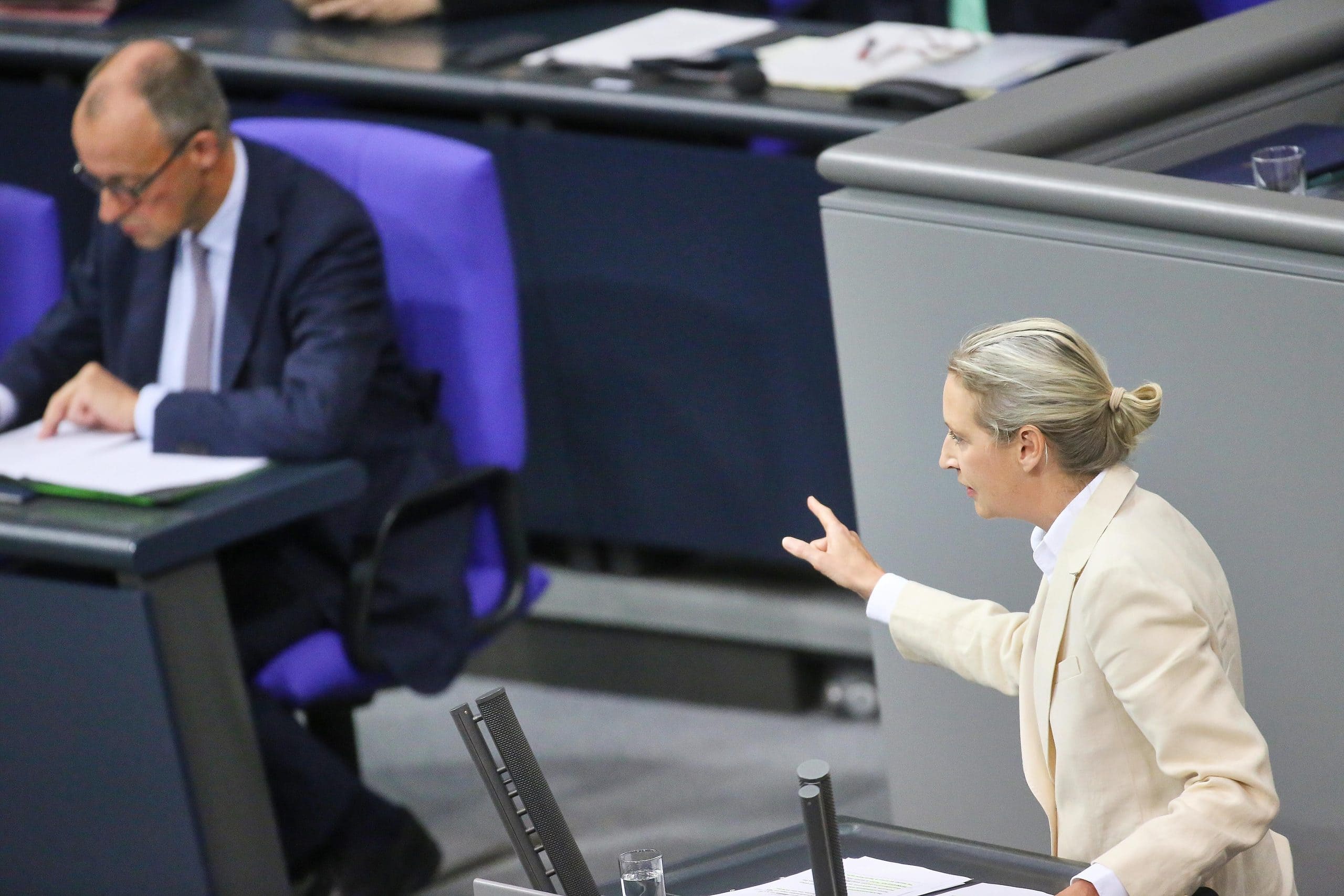 AfD-Chefin Alice Weidel in einem hellen Anzug spricht und gestikuliert an einem Podium, während Bundeskanzler Merz in einem dunklen Anzug an einem nahe gelegenen Schreibtisch sitzt und in einem formellen Rahmen liest.