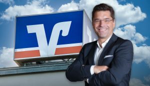 Ein Mann in einem schwarzen Anzug steht lächelnd mit verschränkten Armen vor einem Gebäude mit einem blau-weiß-orangen Bitcoin-Logo.