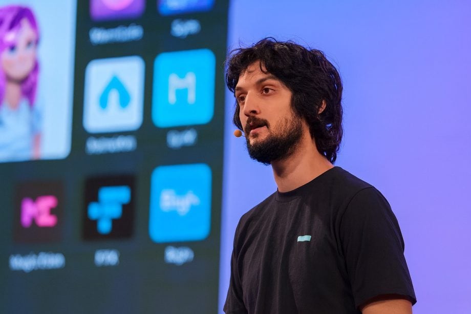 Ein Mann mit einem Headset-Mikrofon steht vor einem Bildschirm, auf dem App-Symbole, Grafiken und Krypto-Logos angezeigt werden, und hält eine Präsentation.