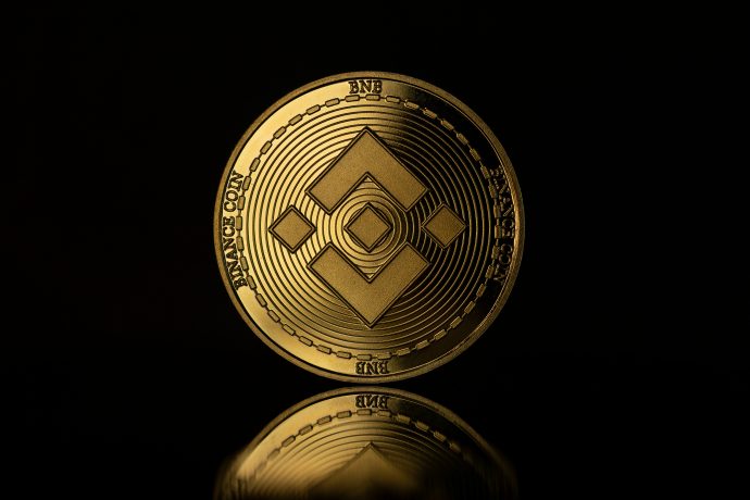 Eine goldfarbene BNB-Münze mit dem Binance-Logo steht aufrecht vor einem dunklen Hintergrund, ihr Bild spiegelt sich auf einer glänzenden Oberfläche.