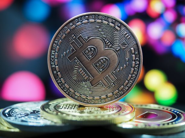 Viertgrößte Währung der Welt: Hat Bitcoin seinen Zenit erreicht? Eine Nahaufnahme einer Bitcoin-Münze, die aufrecht auf mehreren anderen Münzen steht, mit einem bunten, unscharfen Hintergrund.