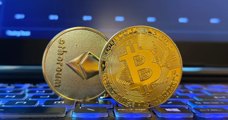 Zwei physische Münzen, die Ethereum und Bitcoin darstellen, stehen auf einer beleuchteten Tastatur, während im Hintergrund auf einem Laptopbildschirm ein Bitcoin-Kurschart mit Allzeithoch aus der aktuellen Handelswoche zu sehen ist.