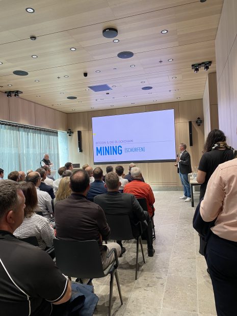 Eine Gruppe von Menschen sitzt in einem modernen Konferenzraum, beschäftigt mit einer BitcoinForum-Sitzung, und hört einem Moderator zu, der vor einem Bildschirm mit dem Titel Bitcoin & die Blockchain Mining (Schürfen) steht.