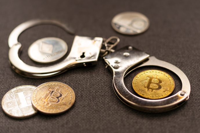 Ein Paar Metallhandschellen und mehrere Kryptowährungsmünzen, darunter Bitcoin und Ethereum, sind auf einer dunklen Fläche ausgestellt und verdeutlichen die Komplexität von Sicherheit und Regulierung im Zusammenhang mit Bitcoin.