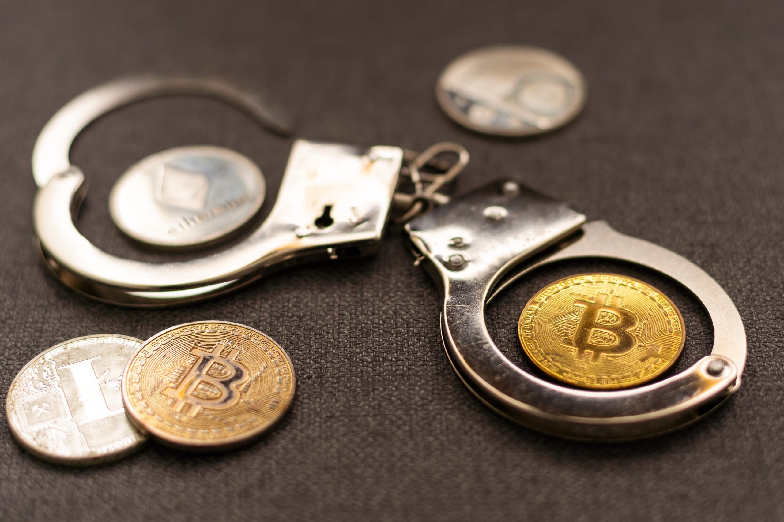 Ein Paar Metallhandschellen und mehrere Kryptowährungsmünzen, darunter Bitcoin und Ethereum, sind auf einer dunklen Fläche ausgestellt und verdeutlichen die Komplexität von Sicherheit und Regulierung im Zusammenhang mit Bitcoin.