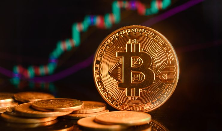 Im Vordergrund ist ein Stapel Bitcoin-Münzen zu sehen, während im unscharfen Hintergrund ein digitales Finanzdiagramm mit Candlestick- und Liniendiagrammen zu sehen ist, die die neuesten Bitcoin-Kurs-Trends veranschaulichen.