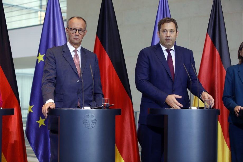 Zwei deutsche Politiker bei einer Pressekonferenz, umgeben von EU- und Deutschlandfahnen – im Kontext wachsender Kritik aus der Bitcoin-Szene am politischen System.