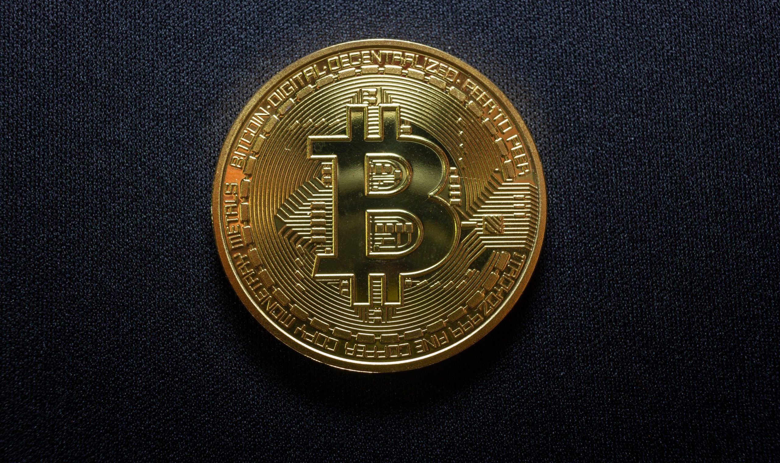 Eine goldfarbene Bitcoin-Münze mit dem Bitcoin-Symbol ruht auf einem schwarzen, strukturierten Hintergrund.