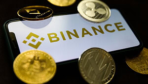 Ein Smartphone mit dem Binance-Logo ist von verschiedenen physischen Kryptowährungsmünzen umgeben, darunter Bitcoin, Ethereum, Litecoin und XRP.