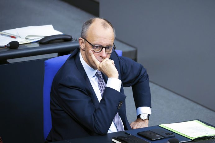 Ein Mann im Anzug und mit Brille sitzt an einem Schreibtisch, die Hand am Kinn und wirkt nachdenklich. Auf dem Schreibtisch liegen Dokumente, ein Telefon und Büromaterialien, wobei ein Bericht über Bitcoin als Reservewährung seine Aufmerksamkeit erregt.