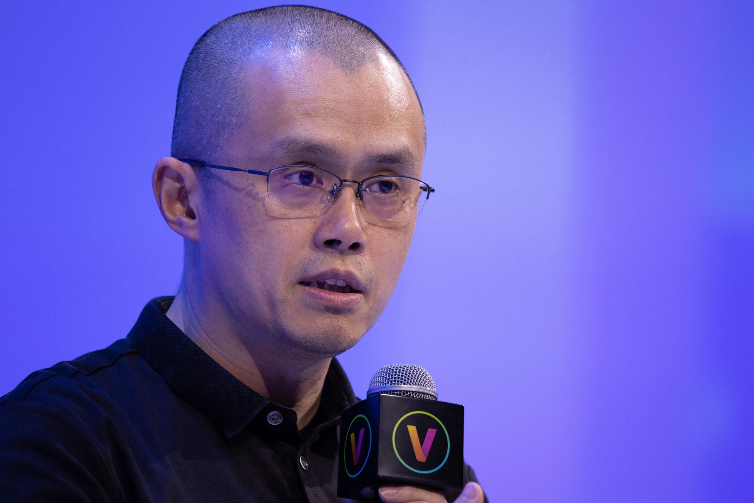 Changpeng Zhao warnt vor CZ-Memecoin: “Don’t buy the meme” Ein Mann mit Brille spricht in ein Mikrofon auf der Bühne vor einem blauen Binance-Hintergrund.