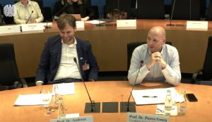 Prof. Dr. Pierre Georg von der Frankfurt School of Finance and Management will die Jahreshaltefrist abschaffen Zwei Männer sitzen an einem Konferenztisch mit Mikrofonen und Dokumenten und diskutieren über Krypto; einer spricht, während der andere zuhört. Das angezeigte Datum ist der 13.10.2025, und im Hintergrund sind weitere Personen zu sehen.