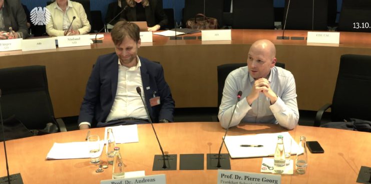 Zwei Männer sitzen an einem Konferenztisch mit Mikrofonen und Dokumenten und diskutieren über Krypto; einer spricht, während der andere zuhört. Das angezeigte Datum ist der 13.10.2025, und im Hintergrund sind weitere Personen zu sehen.