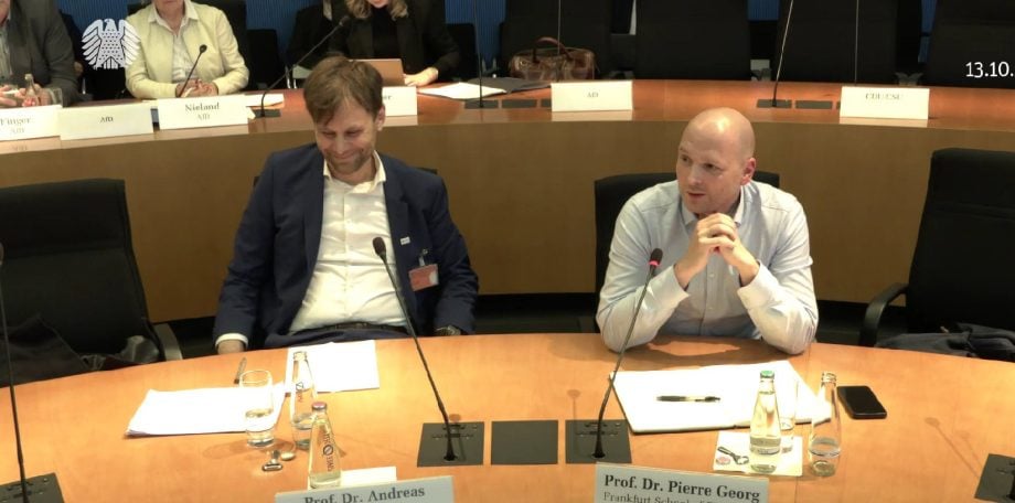 Zwei Männer sitzen an einem Konferenztisch mit Mikrofonen und Dokumenten und diskutieren über Krypto; einer spricht, während der andere zuhört. Das angezeigte Datum ist der 13.10.2025, und im Hintergrund sind weitere Personen zu sehen.