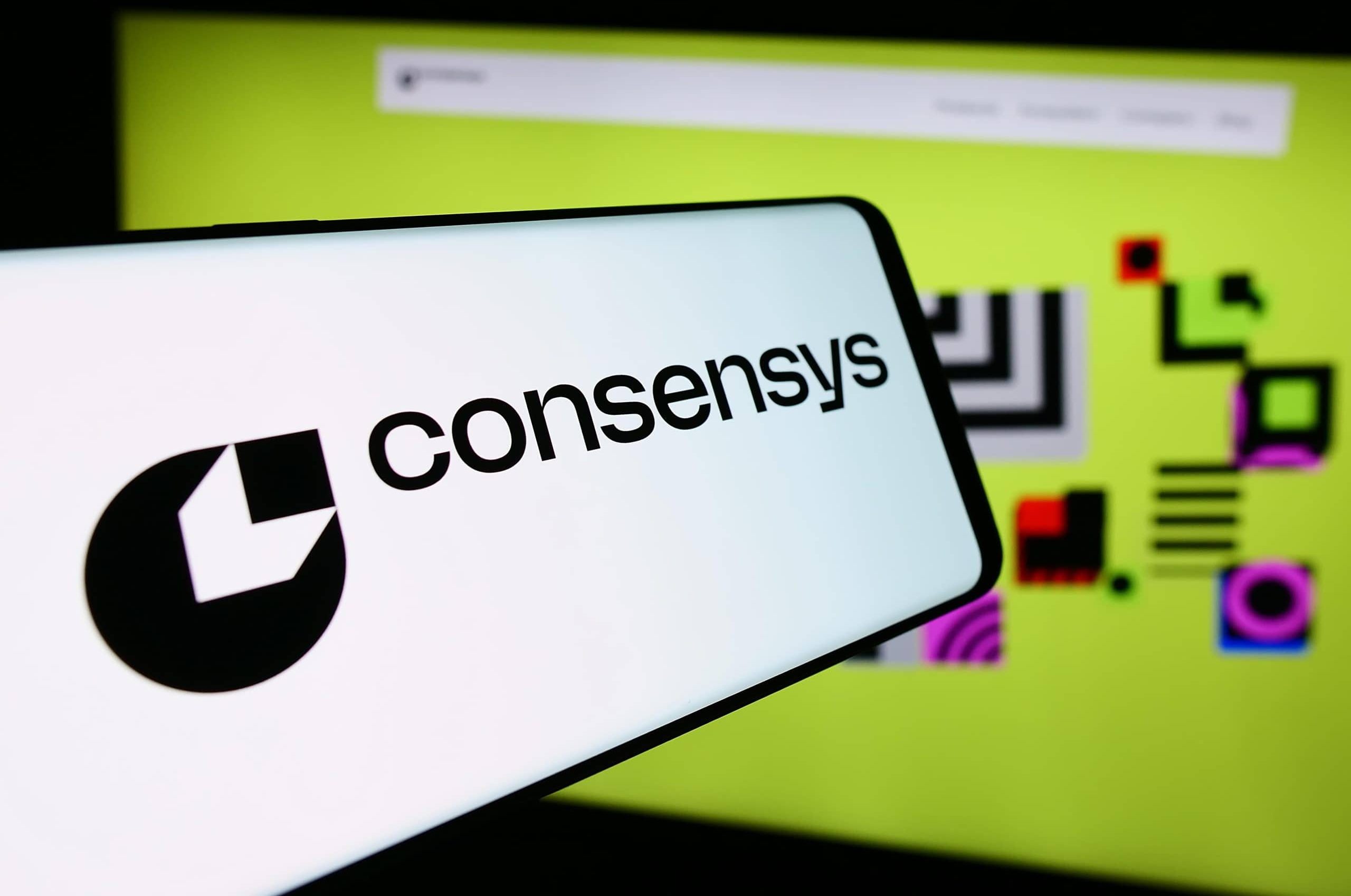 Ein Smartphone zeigt das Logo von ConsenSys, einem führenden Ethereum-Softwareunternehmen, vor einem Computerbildschirm mit abstrakten Formen auf gelbem Hintergrund.