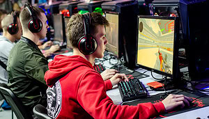 Mehrere junge Leute mit Headsets sitzen an Computern und spielen CS2, ein Ego-Shooter-Videospiel, bei einem esports-Event oder Gaming-Turnier.
