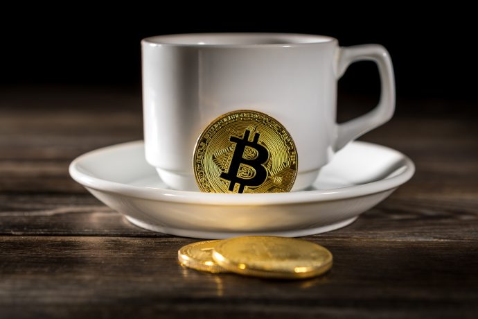 Das sind die 5 wichtigsten Krypto-News der Woche Eine weiße Kaffeetasse und eine Untertasse stehen auf einem Holztisch, eine goldene Krypto-Bitcoin-Münze lehnt an der Tasse und zwei weitere Münzen liegen auf dem Tisch.