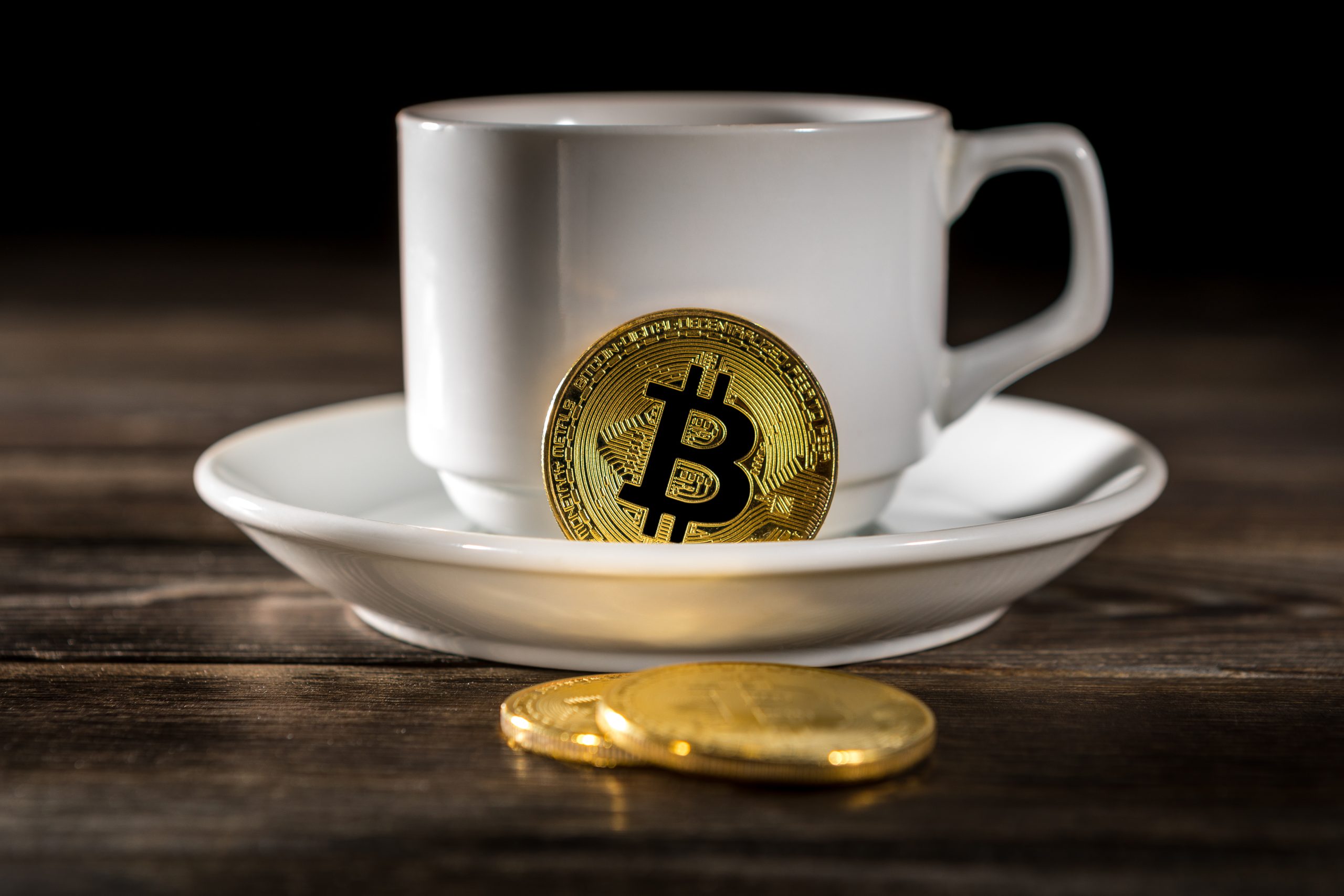 Das sind die 5 wichtigsten Krypto-News der Woche Eine weiße Kaffeetasse und eine Untertasse stehen auf einem Holztisch, eine goldene Krypto-Bitcoin-Münze lehnt an der Tasse und zwei weitere Münzen liegen auf dem Tisch.
