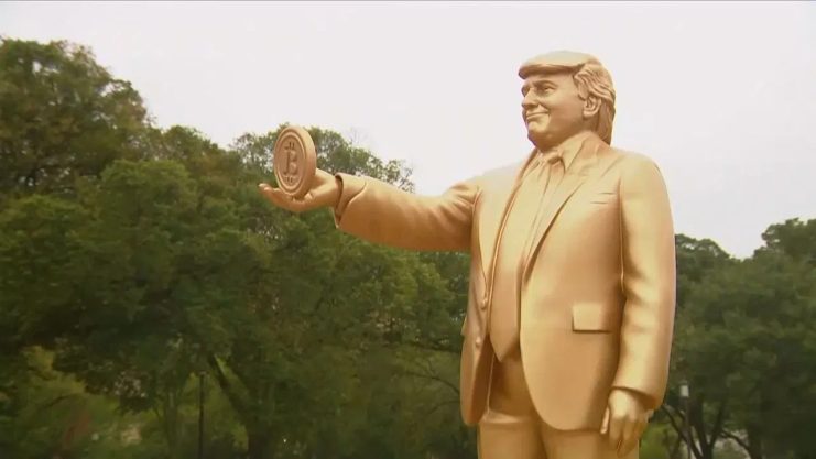 Bitcoin-Erfinder Donald J. Trump? Eine goldfarbene Statue eines Mannes im Anzug, der ein rundes Objekt mit einem Bitcoin-Symbol in der Hand hält - von dem oft behauptet wird, dass es Satoshi oder Trump ähnelt, als ob sie die gleiche Person wären - steht im Freien mit Bäumen im Hintergrund.
