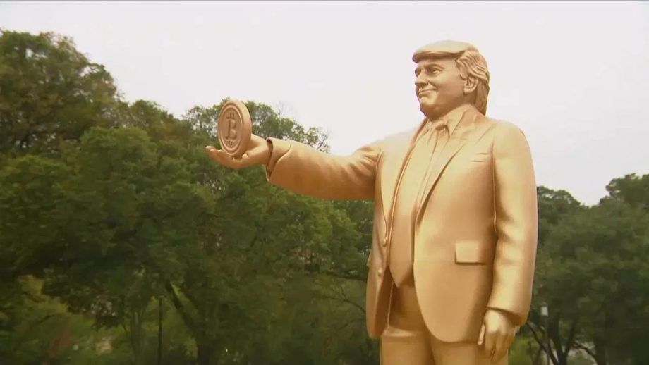 Eine goldfarbene Statue eines Mannes im Anzug, der ein rundes Objekt mit einem Bitcoin-Symbol in der Hand hält - von dem oft behauptet wird, dass es Satoshi oder Trump ähnelt, als ob sie die gleiche Person wären - steht im Freien mit Bäumen im Hintergrund.