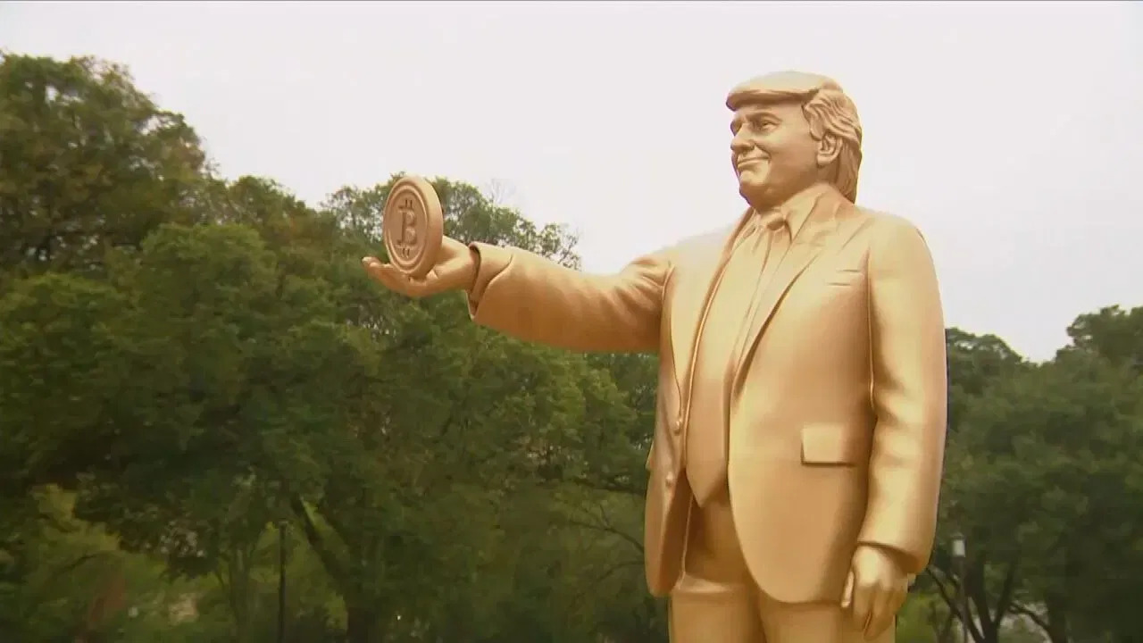 Bitcoin-Erfinder Donald J. Trump? Eine goldfarbene Statue eines Mannes im Anzug, der ein rundes Objekt mit einem Bitcoin-Symbol in der Hand hält - von dem oft behauptet wird, dass es Satoshi oder Trump ähnelt, als ob sie die gleiche Person wären - steht im Freien mit Bäumen im Hintergrund.