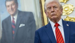 Ein Mann in blauem Anzug und rot gestreifter Krawatte steht vor einem gerahmten Porträt eines anderen Mannes in Anzug und Krawatte, beide strahlen eine Ernsthaftigkeit aus, als würden sie über die Folgen eines kürzlichen Börsen-Crashs nachdenken.