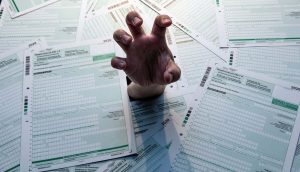 Eine Hand streckt sich durch einen Stapel Steuerformulare, was auf Überforderung oder Stress durch Papierkram hindeutet - selbst wenn man die Komplexität von Krypto nicht berücksichtigt.