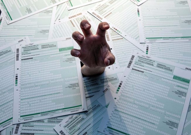 Eine Hand streckt sich durch einen Stapel Steuerformulare, was auf Überforderung oder Stress durch Papierkram hindeutet - selbst wenn man die Komplexität von Krypto nicht berücksichtigt.