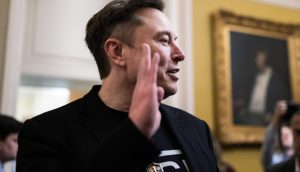 Bitcoin als Antwort auf entgrenzte Geldpolitik Ein Mann in einer schwarzen Jacke hebt seine rechte Hand, während er in einem Haus über Bitcoin spricht. Im Hintergrund sind Menschen und ein gerahmtes Porträt zu sehen.