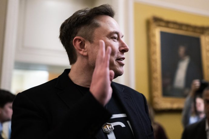 Ein Mann in einer schwarzen Jacke hebt seine rechte Hand, während er in einem Haus über Bitcoin spricht. Im Hintergrund sind Menschen und ein gerahmtes Porträt zu sehen.