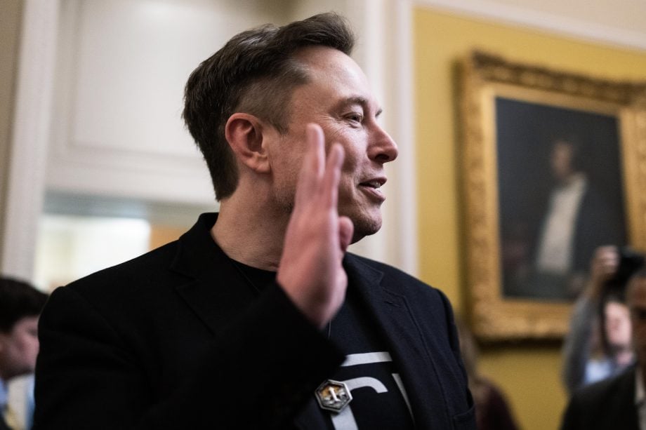 Ein Mann in einer schwarzen Jacke hebt seine rechte Hand, während er in einem Haus über Bitcoin spricht. Im Hintergrund sind Menschen und ein gerahmtes Porträt zu sehen.