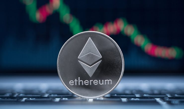 Ethereum muss sich zeitnah stabilisieren Eine silberne Ethereum-Münze liegt auf der Tastatur eines Laptops. Im Hintergrund ist ein unscharfer digitaler Ethereum-Chart mit roten und grünen Kerzen zu sehen.
