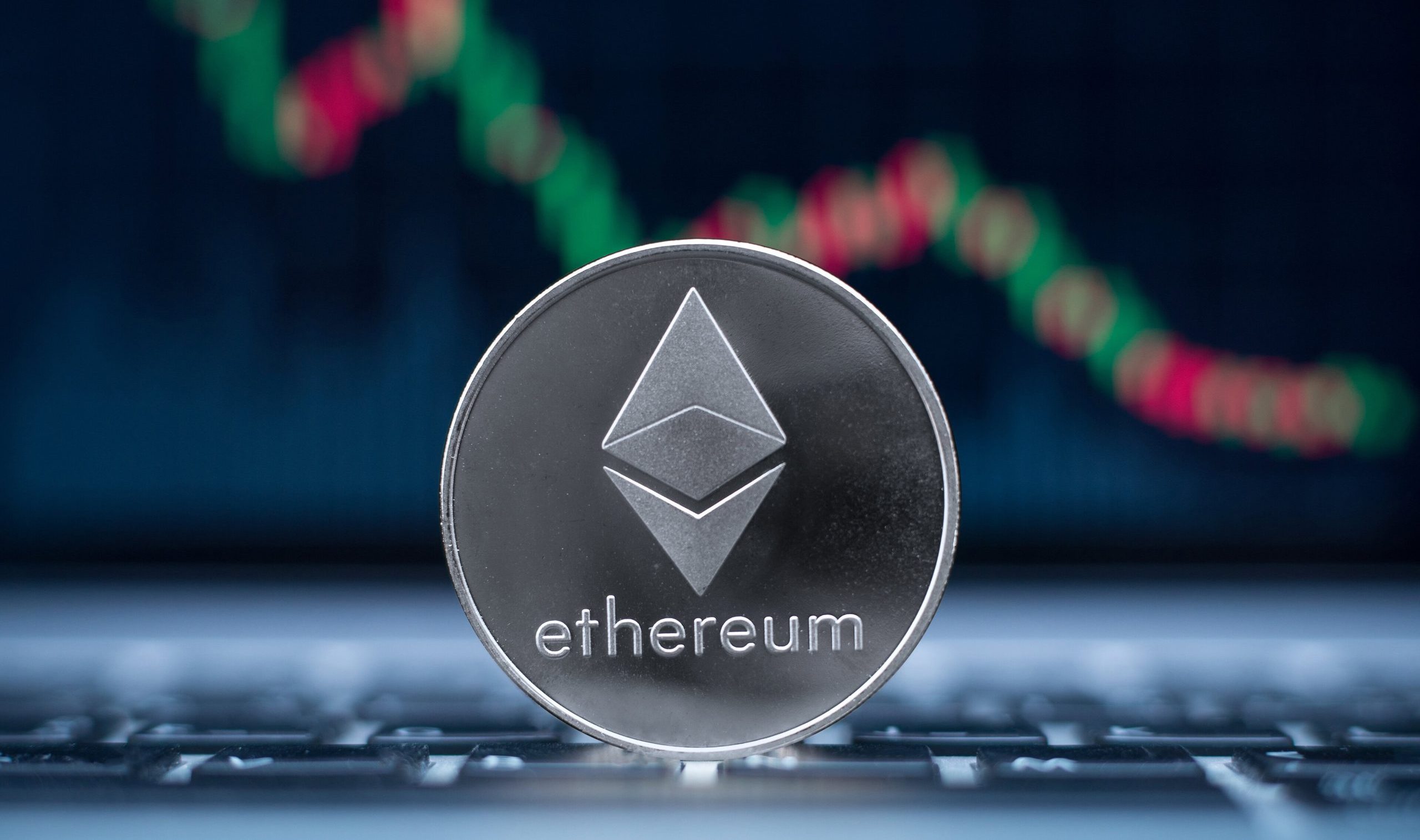 Ethereum muss sich zeitnah stabilisieren Eine silberne Ethereum-Münze liegt auf der Tastatur eines Laptops. Im Hintergrund ist ein unscharfer digitaler Ethereum-Chart mit roten und grünen Kerzen zu sehen.