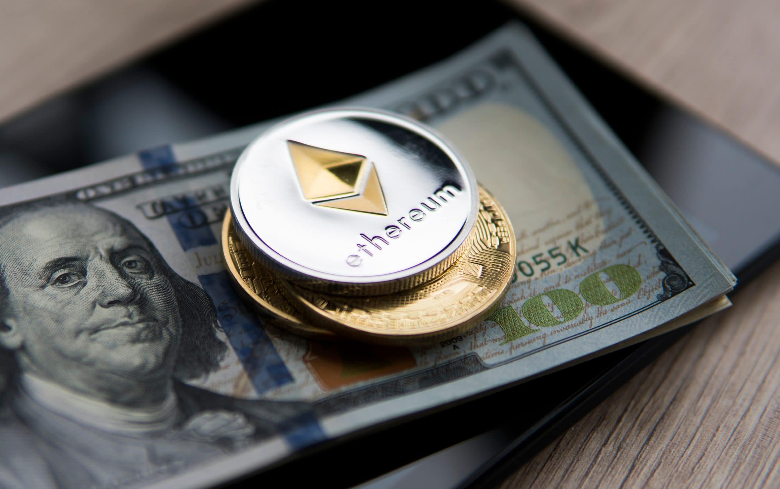 ethereum-treasury-bitmine-14-milliarden-usd Eine silberne Ethereum-Münze und zwei Goldmünzen liegen auf einem 100-US-Dollar-Schein, der auf einem schwarzen Smartphone auf einer hölzernen Oberfläche platziert ist - ein Symbol für digitale Vermögenswerte wie BitMine in der modernen Finanzlandschaft.