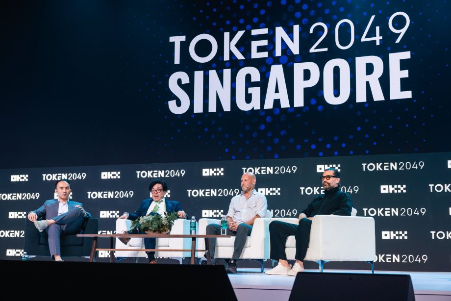 Vier Männer, darunter Sam Tabar, sitzen während einer Podiumsdiskussion auf der TOKEN2049-Veranstaltung in Singapur auf einer Bühne, während im Hintergrund das Branding der Veranstaltung und Gespräche über Bitcoin zu sehen sind.