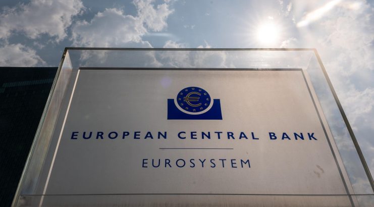 Schild für die Europäische Zentralbank mit dem Logo und dem Wort Eurosystem, fotografiert im Freien bei teilweise bewölktem Himmel mit Sonnenlicht im Hintergrund.
