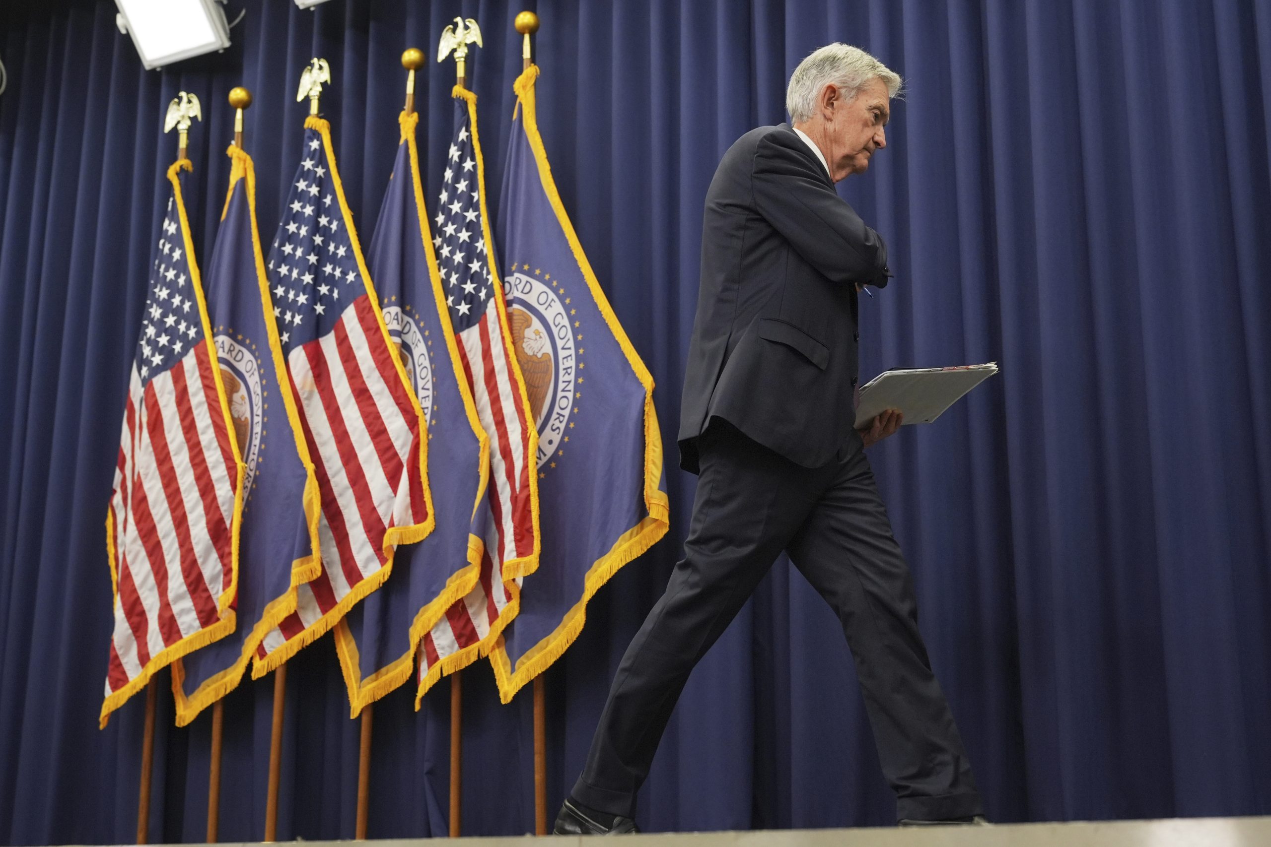 fed-chef-5-kandidaten Ein Mann im Anzug geht über die Bühne und hält eine Mappe in der Hand. Im Hintergrund sind mehrere Flaggen der USA und der Federal Reserve zu sehen - möglicherweise einer von Powells Nachfolge-Kandidaten.