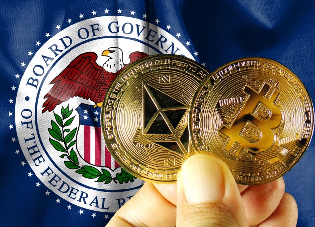 Eine Person hält Ethereum- und Bitcoin-Münzen mit der Aufschrift "Krypto" vor einem Emblem der US-Notenbank.