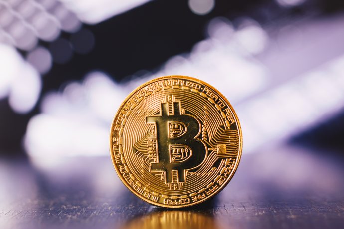Ein goldfarbener Bitcoin mit dem ikonischen Bitcoin-Logo steht aufrecht auf einer spiegelnden Oberfläche mit einem unscharfen Hintergrund.