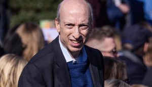 Gary Gensler, ein glatzköpfiger Mann in einem dunklen Blazer und einem blauen Pullover steht im Freien inmitten einer Menschenmenge, mit etwas Grün im Hintergrund, möglicherweise bei einer Veranstaltung von Gensler.