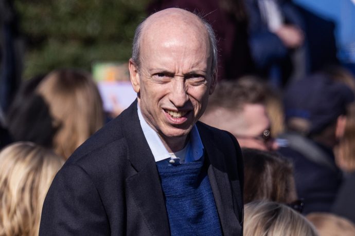 Gary Gensler, ein glatzköpfiger Mann in einem dunklen Blazer und einem blauen Pullover steht im Freien inmitten einer Menschenmenge, mit etwas Grün im Hintergrund, möglicherweise bei einer Veranstaltung von Gensler.