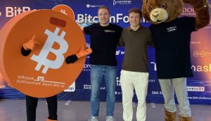 Drei Personen posieren vor einer BitcoinForum-Kulisse: eine trägt ein Bitcoin-Münzen-Kostüm, eine andere ist leger gekleidet, und die dritte trägt ein Bärenkostüm.