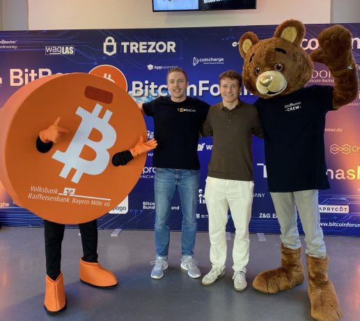 Drei Personen posieren vor einer BitcoinForum-Kulisse: eine trägt ein Bitcoin-Münzen-Kostüm, eine andere ist leger gekleidet, und die dritte trägt ein Bärenkostüm.