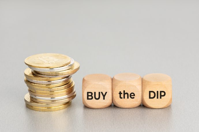 Das Bild zeigt Bitcoin-Münzen neben Würfeln auf denen "Buy the DIP" draufsteht