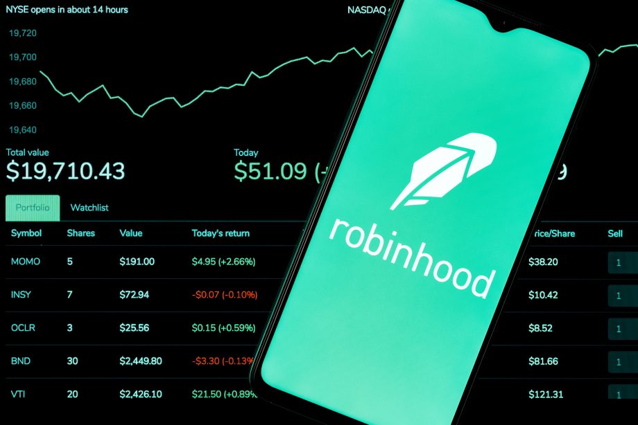 Ein Smartphone zeigt das Logo der Robinhood-App vor einem Computerbildschirm mit Börsendaten und einem steigenden Robinhood-Liniendiagramm.